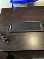 Solar panel displayed on a wooden surface, showing full front view, black plastic casing, and attached cable.