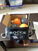 Open black fabric carrying bag containing four bocce balls (2 orange, 2 dark gray) and white pallino ball on table.