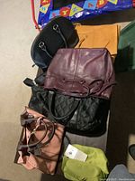 Photo showing 7 purses stacked or arranged including pink handbag with brown handles, black quilted bag, maroon drawstring purse, and others