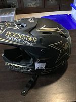 Side view of the medium size Rockstar Kinetic helmet showing black matte finish with gold Rockstar Energy Drink logos and stars.