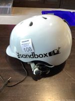 Side view of white Sandbox helmet with black logo and visible attached audio cable with 3.5mm jack; minor scuff marks on surface.