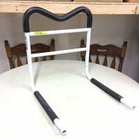 White metal bed rail with V-shaped black padded top handle and black padded lower legs, shown from front angle on white surface with wooden chairs in background.