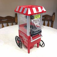 Front angled view showing full popcorn maker with red and white striped roof, clear viewing panels, black wheels and front logo panel.