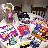 Full view of the collection including the Rapunzel Disney Animators Collection doll in box, Twilight book, Big Bang Theory DVD, Speak Out game, Pictionary game, card game, and two cartoon-themed belts on table.