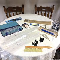 Photo showing an assortment of vintage drafting tools arranged on a white table including flexible curves in fabric case, wooden and plastic rulers, metal set squares, compass, drawing brush, magnifier, and pens.