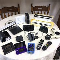 Overall view of various electronic devices arranged on a white tablecloth: gaming consoles, routers, Apple TV, phone, hard drive, hair dryer, remote, and cords.