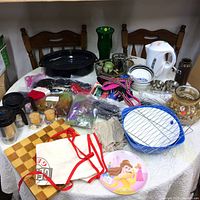 Wide view of various household items on a table including a white kettle, blue roasting pan, wooden cutting board, aprons, dog harnesses, yarn, travel mugs, and miscellaneous small kitchen and decorative items.