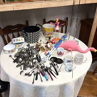 Photo of entire table showing assorted cutlery, mugs, popcorn bucket, pink flamingo decor, plush toys, and other household items.