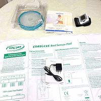 Photo of the bed sensor pad laying flat showing printed instructions and charger cable with plug. Digital blood pressure monitor with cuff visible to the top right. Clear plastic cast protector with blue seal in original packaging also visible.