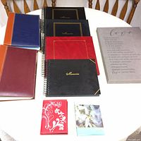Collection of multiple photo albums of different colors and sizes arranged on white table with a beige canvas featuring a love poem printed in script.