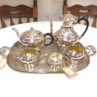Full view of the tea and coffee service on a tray, showing the set of silver-toned pots and bowls with black handles on the pots and a large serving tray beneath.