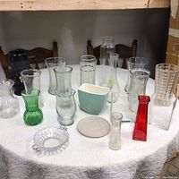 Wide view of fourteen glass and ceramic vases of various shapes and colors arrayed on white cloth surface, mostly clear glass with colorful accents.
