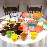 Full set of assorted vintage melamine cups, bowls, mugs, plates, and cookbooks displayed on table with chairs in background.