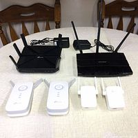 Photo showing all six TP-Link networking devices: 2 black routers and 4 white WiFi extenders on a table.