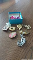 Photo showing eleven assorted brooches in various designs and materials arranged on a wooden surface with a blue box containing two fabric flower brooches.