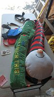 Two textured large plush crinkle dog toys, one green with multi-color buttons and KONG text, other red and green striped with a smiling elf/gnome face, resting on a table with leashes and coat.