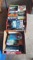 Two boxes filled with a variety of hard and soft covered fiction books stacked tightly. Visible titles include James Patterson's The Cross and Iain Banks' The Bridge.