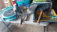 Table with rackets including squash, tennis, and kids rackets; croquet mallets and wickets; Ogosport sports disk; boxed Black Diamond Racer snow slider; Slip 'n Slide packaging partly shown