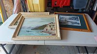 Two framed paintings and one framed print placed on a white table. One painting shows a coastal landscape with water and house. Print is dark and white with a brown frame.
