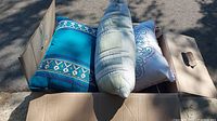 Opened cardboard box containing several throw pillows with varied colors and designs outdoors in daylight.