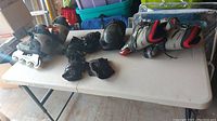 Two pairs of roller blades and protective pads displayed on a white folding table. Includes black elbow pad, multiple black knee pads and wrist guards, and two pairs of roller blades.