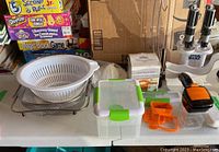 White plastic strainer with attached pan, foldable legs, container with green lid, orange cutters, and boxed Pampered Chef juicer alongside Star Wars popsicle maker and banana holder.