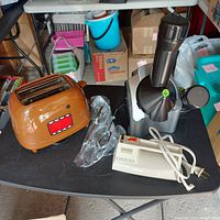 Three kitchen appliances on black table: brown toaster with cartoon face, white GE hand mixer with beaters, silver-black Yonanas yogurt maker.