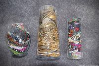 Photo showing three large clear jars filled with assorted jewelry including colorful beaded pieces and gold-tone chains