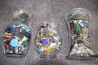 Three large glass jars packed full of assorted jewelry including necklaces, bracelets, and beads in various colors and materials, shown from front view.
