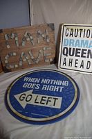 Three wood décor signs arranged together: one 'Man Cave' sign made with rocks on wood with metal corner braces, one 'Caution Drama Queen Ahead' sign with visible water damage, and one round blue 'When Nothing Goes Right Go Left' sign.