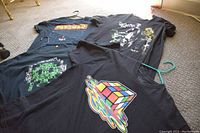 Four black medium t-shirts laid on carpet floor with visible colorful vintage graphic prints including a melting Rubik's Cube, Pac-Man maze, Zelda characters, and pixel art design.