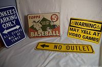 Four vintage metal signs displayed together (Yankees parking, Topps baseball card, video games warning, no outlet).