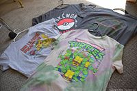 Four vintage graphic T-shirts laid out on carpet with hangers visible, two featuring Pokemon, two Teenage Mutant Ninja Turtles, one partially obscured Big League Baseball shirt visible.