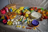 Wide assortment of colorful plastic food items and kitchenware arranged on a light surface showing picnic basket, plates, plastic fruits and vegetables, eggs, utensils and more.