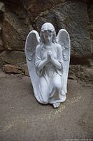 Front view of white kneeling winged angel statue on ground with rock background