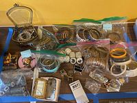 Wide view of lot showing jewelry box, multiple sealed bags of bangles, brooches and earrings.