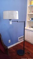 Full view of the tall floor lamp with white drum shade against a blue wall and hardwood floor, showing extendable arm and round base.