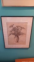Framed wall art showing detailed pencil or ink drawing of flowers in a vase hung on a teal wall.