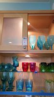 Shelf of assorted colored and frosted glassware including teal goblets and red drinking glasses