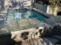 Stack of clear square glass block tiles of various sizes placed on stone ledge by pool. Colors appear clear with weathered, slightly dirty surfaces.