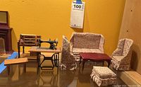 Wide shot showing the entire miniature set including sofa, chairs, ottoman, coffee table, and sewing machine set against a yellow wall and ruler.