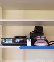 Three items on shelf: black RCA VHS recorder, Polaroid Supercolor 635CL instant camera, rock tumbler motorcycle helmet style with red control buttons