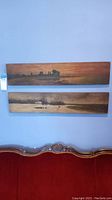 Two long horizontal wooden panel paintings with landscape scenes hung on a blue wall above a decorated red couch.