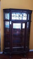 Front view of vintage wooden curio cabinet showing decorative leaded glass door and curved glass panels.