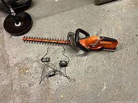 Full view of the Black + Decker cordless hedge trimmer and its charger cords lying on a concrete floor.