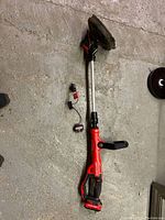 Full view of the red Craftsman weed wacker showing shaft, handle, battery pack, and trimming head on a concrete floor.
