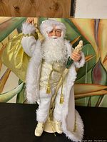 Full body view of a large Santa ornament in white and gold holding a gold sack and tree with a stick