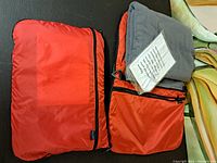 Orange nylon zippered carrying cases containing travel pillows, one larger rectangular and one smaller pouch with visible gray pillow inside