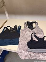 Four women's sports shirts laid out on a surface: black and blue sports shirt with mesh panel, sleeveless spotted pattern tank top, black racerback sports bra, and black and gray Lululemon sports bra.