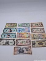 Top view photo showing 12 currency notes laid out in rows, including novelty Disney notes and Japanese banknotes with various portraits and denominations.